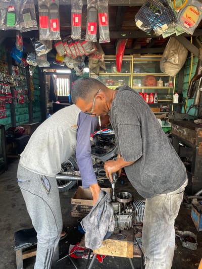 The image captures a bustling motorcycle repair shop in Glee Gogo, Indonesia, with two men focused on their work. In the foreground, a man wearing a grey t-shirt with blue long sleeves, grey sweatpants, and glasses observes or assists, with a cigarette in his mouth. His shirt bears the text "Arada Jeans." Behind him, another man in a dark grey t-shirt and light grey pants is actively engaged in repairing a disassembled motorcycle engine, holding a tool. The engine is positioned on a wooden block on the dark, utilitarian floor.
The shop environment is typical of a local garage, filled with various motorcycle parts and accessories. These items are stored on wooden shelves and hung around the space. Visible products include packaged drive belts, air filters, and bottles of engine oil. Text such as "MACHO," "YAMAHA," and "HONDA" can be seen on different packages. A cardboard box labeled "Eralube 2TR" is also present. The walls are a mix of wood and green corrugated material. The scene is illuminated by ambient daylight, indicating it is daytime. The overall atmosphere is one of practical, hands-on repair.