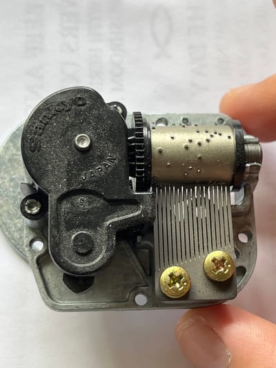 The image presents a close-up, top-down view of a music box mechanism, held by two human fingers partially visible at the top of the frame. The central component is a golden-colored metal cylinder covered with numerous small pins that project outwards. Below the cylinder is a black gear. Adjacent to the cylinder is a metal comb composed of multiple tines of varying lengths, which would produce musical notes when struck by the cylinder's pins. This comb is secured to a grey metal base plate by two brass screws with Phillips head drives. A prominent black plastic housing, likely encasing the winding mechanism and gears, occupies the lower part of the mechanism. The words "Sankyo" and "JAPAN" are clearly embossed on this black housing. The mechanism appears clean and well-preserved. The background is a white surface, possibly a piece of paper, featuring faint, reversed text and symbols, including fragments like "HE," "DW," "MOON," "MIND," "AA," "EEL," and a stylized fish outline. The overall impression is an inspection or detailed presentation of the music box component.
