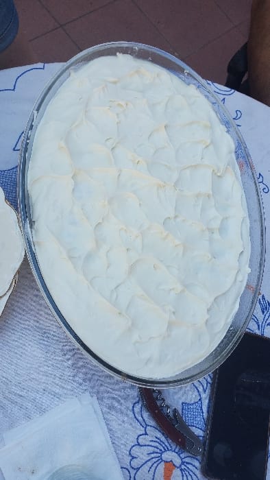 The scene depicts an overhead view of a large, oval-shaped clear glass dish filled with a light, textured dessert, likely a cream or meringue, displaying a rippled surface. The dish is placed on a light-colored tablecloth patterned with blue and orange floral designs. To the lower left of the dish, a folded white paper napkin is visible. To the lower right, a corkscrew with a dark red handle and silver metal parts lies next to a black smartphone, screen facing up. The background reveals a reddish-brown tiled floor with visible grout lines in the upper left, and a human leg clad in dark fabric in the upper right, indicating the presence of an individual. The setting is located in the city of Partido de Quilmes, Argentina.