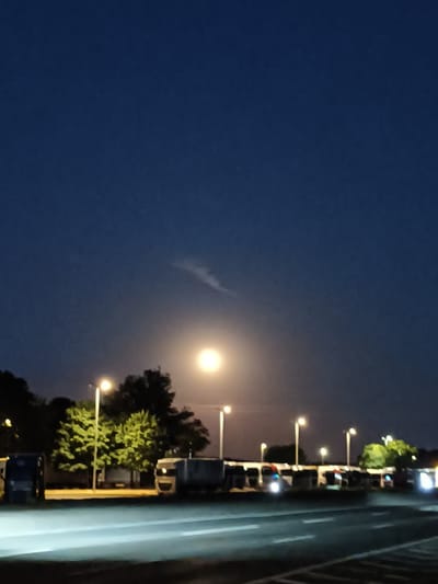 The image depicts a night scene, possibly a parking lot or rest stop near a road, in Münster, Germany. The sky is a deep, dark blue, with a bright, hazy moon positioned in the upper middle portion of the frame. A faint, wispy cloud formation is visible near the moon. In the middle ground, several trees with green foliage are illuminated by artificial lights. A row of parked vehicles, including what appear to be trucks or commercial vans, are visible. Streetlights with yellow illumination dot the landscape. In the foreground, a road or paved surface is present, reflecting the light from the vehicles and streetlights, creating streaks of white light. The overall impression is one of a quiet, nocturnal setting on the outskirts of a city.