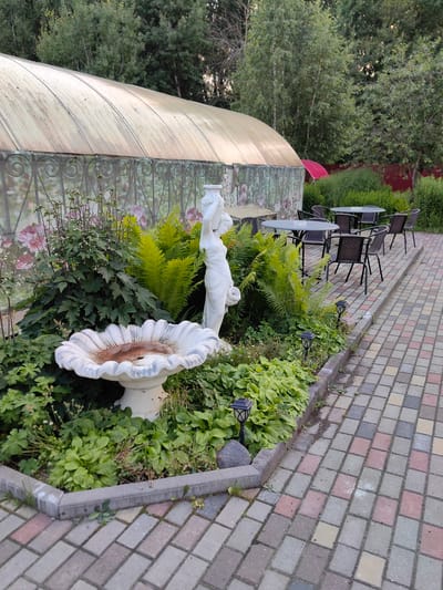 In the city of Фаніпальскі сельскі Савет, Belarus, an outdoor seating area is depicted. A patterned brick walkway lines the foreground, leading towards several round tables with dark chairs. Behind the tables, a garden bed features a white, ornate birdbath as its centerpiece, surrounded by lush green foliage, ferns, and a white statue of a woman. A greenhouse with a translucent, curved roof stands in the background, partially obscured by trees and a red umbrella. The scene is set during daylight hours, with natural light illuminating the area.