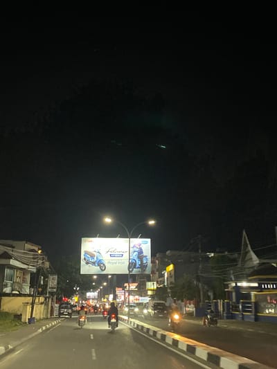This is a night time photograph of a street in Walennae, Indonesia. The street is paved and has multiple lanes of traffic. There are several vehicles on the road, including cars and motorcycles. The buildings on either side of the street are illuminated, and there is a large billboard advertising Yamaha scooters. The sky is dark and clear, with some faint green lights visible in the distance. The overall atmosphere is busy and urban.