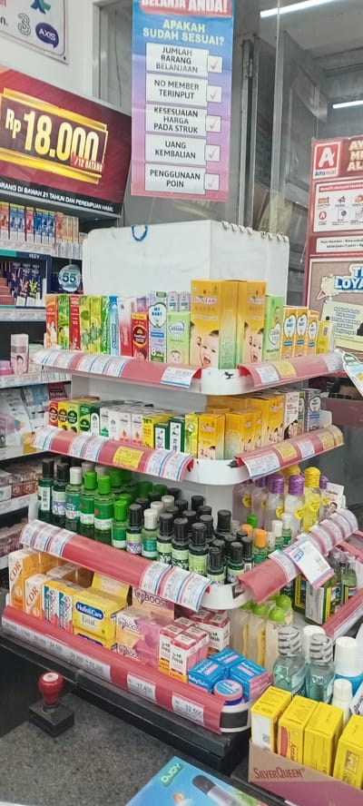 The photograph, taken at eye-level, shows a close-up view of a retail shelf in what appears to be a store, likely in Payakumbuh, Indonesia based on the Indonesian language on the signs. The shelf is stocked with a variety of personal care and health-related products, including baby oil, cough medicine, vitamins (such as CDR and Imboost), and mouthwash (Listerine). The items are arranged in rows and columns, with some products stacked on top of others. Red price strips displaying prices in Indonesian Rupiah (Rp) run along the front of the shelves.

Several promotional signs are visible. One sign advertises a product for Rp 18,000 for 12 pieces and warns against usage by individuals under 21 and pregnant women. Another lists criteria for a satisfactory purchase, including correct item count, no member input required, correct pricing, correct change, and point usage. A sign is for Alfamidi, a grocery store chain. There are signs showing words like "axis", "veeV", and "Play", seemingly logos of different companies.