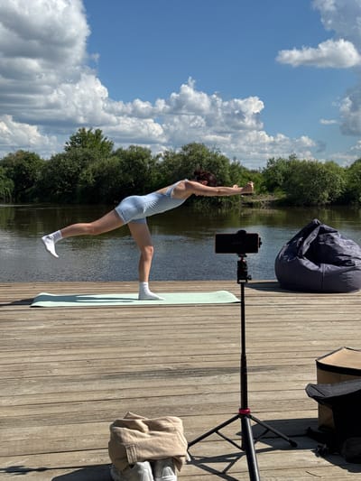 On a wooden deck by a body of water, a woman performs a yoga pose. She is wearing light blue athletic wear and white socks, balancing on one leg with her arms extended forward. A light green yoga mat is placed beneath her. To the right of the yoga mat, a black tripod holds a smartphone, facing the woman. Further to the right, a dark gray beanbag chair is positioned. In the foreground, a beige sweatshirt is draped over a pair of light gray sneakers. In the background, lush green trees line the opposite bank of the water, under a bright blue sky with scattered white clouds. The location is identified as the city of Onochino, Russia.