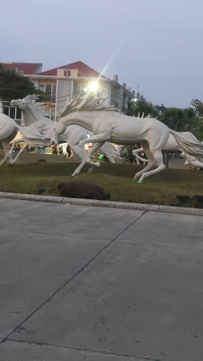 In the city of Pusakarakyat, Indonesia, a group of white horse sculptures gallop across a grassy area. The sculptures are depicted in mid-stride, with their manes and tails flowing dynamically. Behind the horses, a building with a red roof and multiple stories is visible, along with some trees and other foliage. The sky above is overcast, suggesting it is either early morning or late afternoon. In the foreground, a paved area with distinct lines and markings indicates a road or pathway. A dark, oblong object, possibly a large rock or a piece of debris, rests on the grass in front of the sculptures. Several people are present in the background, near the building, with some appearing to be on motorcycles.
