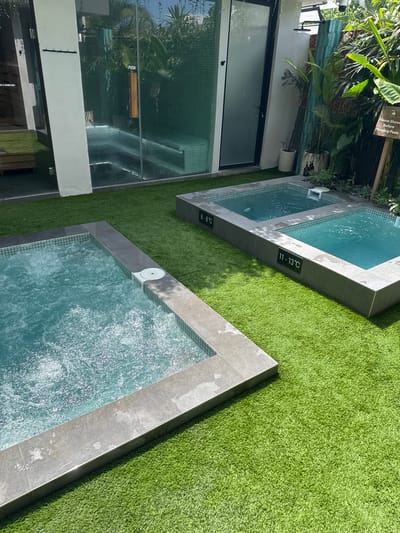 The image shows two plunge pools situated outdoors in Pererenan, Indonesia. The pools are bordered by gray tiles and surrounded by green artificial turf. One pool has a temperature reading of 11-13°C, while the other pool reads 6-8°C. A building with glass doors and windows is visible in the background, along with tropical plants. The scene appears to be a spa or wellness facility.