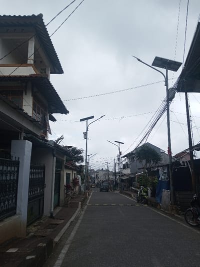 A gray asphalt road stretches into the distance, lined with houses and streetlights. Overhead, numerous power lines crisscross the sky. Several parked motorcycles and a dark-colored vehicle are visible along the sides of the road. The buildings vary in style, some with traditional tiled roofs and others with more modern designs. The sky is overcast, casting a muted light over the scene. A speed bump runs across the road.