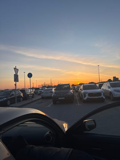 The image depicts a parking lot in Bolshoye Savino, Russia, at sunset. A row of cars is parked diagonally along the right side of the frame, with the open door of a car on the right side dominating the foreground. The sunset casts an orange glow in the background, between cars and poles. Road signs are visible in the middle of the frame. The sky is blue near the top.