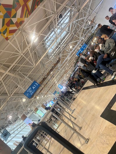 The photo depicts the interior of an airport terminal, possibly Bole International Airport, featuring a complex network of metal support beams forming the roof structure. A sign indicating "Gate A13" is visible, with text in English and an East Asian language. Seated passengers are present in the waiting area. Additional signage displays directions for gates A1-A12, duty-free shop, prayer room, and gates B1-B7. Some people seem to be in line.