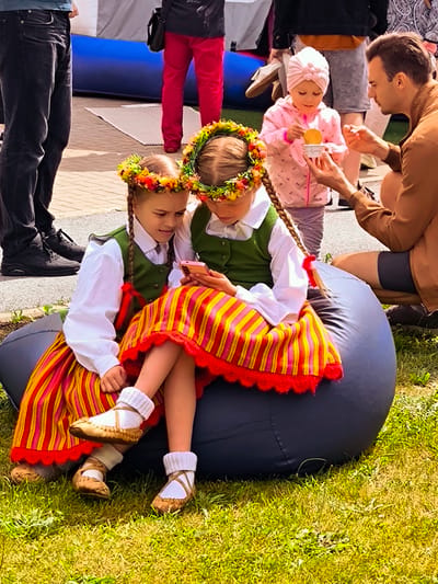 This image depicts an outdoor daytime scene at what appears to be a public gathering or festival in Latvia. No specific architectural or textual cues within the image definitively identify a precise location within the city of Rīga.
In the foreground, two young girls, estimated to be between 6 and 10 years old, are seated on a dark grey, inflated beanbag positioned on a grassy area. Both girls are dressed in traditional Latvian folk costumes, featuring long-sleeved white blouses, green vests, and long, horizontally striped skirts in hues of red, yellow, and orange, trimmed with red lace. Each girl wears a floral wreath (vainags) on her head and has long braids, one of which is tied with a red ribbon. Their footwear consists of white knitted socks and light-brown, traditional-style leather sandals or moccasins. The girl on the right holds a pink smartphone, which both girls are observing intently, suggesting a shared activity.
In the mid-ground, an adult male, likely in his 30s or 40s, is crouching. He wears a brown collared shirt and dark shorts. He extends his left hand towards a small child, approximately 2-4 years old, who is dressed in a pink hooded jacket with a subtle pattern, light-colored pants, and a pink turban-style headwrap. The child holds a small white cup containing a circular, yellow food item, possibly a cookie or waffle, and appears to be interacting with the man.
The background displays additional individuals and elements typical of an event. Partially visible figures include an adult wearing dark jeans and black sneakers on the far left, and another individual in a dark top and bright red pants carrying a black shoulder bag behind them. A large blue inflatable structure and parts of white tent-like structures are also visible. The ground consists of green grass in the immediate foreground, transitioning to a paved surface, likely concrete or interlocking pavers, towards the mid-ground and background. The scene is brightly illuminated by natural sunlight.