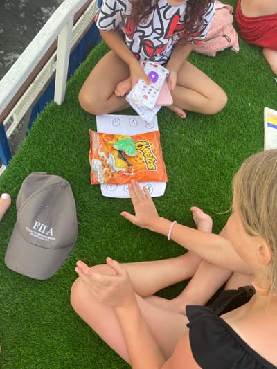 The image shows two young girls sitting on artificial grass, likely outdoors. One girl on the left is wearing a colorful t-shirt with a black and red abstract pattern and has dark curly hair. She is holding a pink object and a white pouch with colorful buttons and drawings. The other girl on the right has blonde hair and is wearing a black ruffled top. She is wearing a pink and white braided bracelet and has her legs crossed.
Between the girls is a bag of Cheetos snacks. Next to the Cheetos, there is a white piece of paper with some drawings and what appear to be simple math symbols like a plus sign and a less than sign. To the right of the Cheetos, a grey Fila baseball cap with "FILA ITALIAN SPORTSWEAR BRAND ESTABLISHED IN 1911" printed on it is lying on the grass. A person wearing red shorts is partially visible in the upper left corner.
The setting suggests a casual outdoor gathering, possibly a picnic or a playtime session. The bright lighting implies it is daytime, and the overall mood appears relaxed and playful. The presence of snacks and drawing materials indicates a leisurely activity.