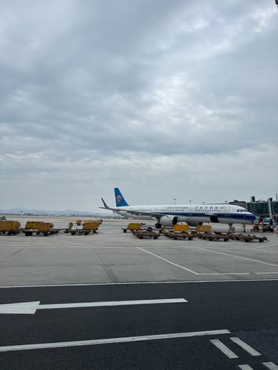 The image shows a China Southern Airlines aircraft parked on the tarmac at Guangzhou Baiyun International Airport in Baiyun District, China. The sky is overcast. The aircraft is connected to a passenger boarding bridge. Ground service equipment is staged nearby. Markings are visible on the tarmac.