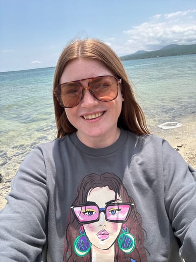 A fair-skinned woman with auburn hair and freckles is taking a selfie at the beach. She is wearing large, amber-tinted aviator sunglasses and a gray crewneck sweatshirt with a graphic print of a woman with pink sunglasses and large green and purple earrings. The background shows a clear blue sky, calm turquoise water, a sandy beach with small rocks, and distant green mountains. The location is not Tsamakaberd, Armenia; it is a coastal area.