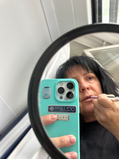 A woman is taking a selfie in a mirror, applying lipstick with a makeup pencil. She is holding a light blue phone with stickers on the back, one a rectangular “Ocean Beach” sticker and another that looks like an airline boarding pass. The mirror appears to be round and is placed against a white surface. The environment is indoors, possibly a bathroom or a similar space with white walls and a window in the background. There is no indication of the location being in Dnp Ryzhiki, Russia.