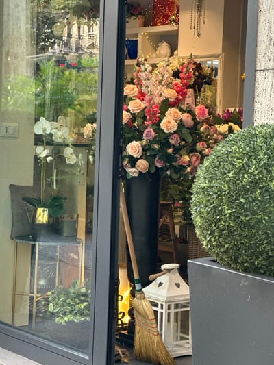 A floristry shop window in Trieste, Italy, displays a large bouquet of pink and cream roses and snapdragons, a tall black vase, a white lantern with heart cutouts, and a broom. To the left, through the glass, a white orchid plant in a gold pot sits on a marble-topped stand next to a candle. Behind the orchid, a digital thermometer displays 25.2 degrees Celsius and 25 degrees Fahrenheit. The exterior facade of a building with textured stonework is visible to the right.