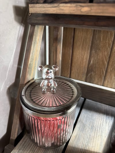 A small, shiny, silver-colored bear figurine is perched on the lid of a clear, fluted glass jar. The lid has a ribbed pattern radiating from the center. Inside the jar, pink objects are visible. The jar sits on a wooden surface, and behind it, a wooden structure with vertical slats is visible. To the left, a textured, light-colored wall with shadows cast upon it is present. The overall scene is indoors, with artificial lighting. The location is not specified in the image, nor is there any visual information that could indicate it is in Семилуки, Russia.