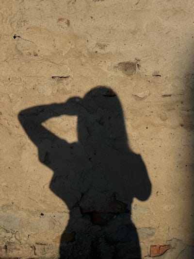 A shadow of a person is cast on a weathered, light brown wall. The shadow depicts a person from the shoulders up, with one arm raised and bent at the elbow, hand touching the back of the head. The person's hair appears to be long, falling down their back. The wall has visible cracks, rough textures, and some exposed brickwork in the lower section. The lighting suggests it is daytime, with a strong light source creating the shadow. The environment is an outdoor setting, likely in Ilavche, Ukraine, given the architectural style of the wall.