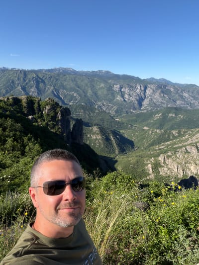 A man with short grey hair, stubble, and sunglasses takes a selfie outdoors. He is wearing a green t-shirt. Behind him, a lush green mountainous landscape stretches under a clear blue sky. The mountains are covered in dense vegetation, with rugged rock formations visible in places. The foreground is filled with green foliage and some yellow wildflowers. The location is Kućišta, Montenegro.