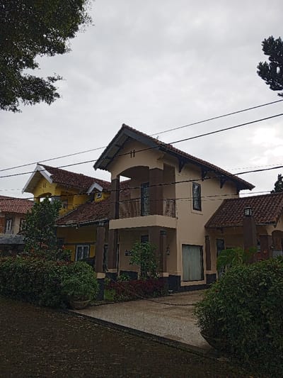 The media file captures a residential scene in Phoukhoun, Laos, featuring several multi-story houses under an overcast, light grey sky. The dominant structure is a two-story house in the foreground, colored beige or cream with dark brown accents on pillars, window frames, and roof eaves. It features a red-brown tiled roof with a pronounced A-frame gable over the second-story balcony. This balcony has a black wrought-iron railing, and the ground floor displays a large window or door with light-colored curtains. Small decorative supports are visible under the roof eaves. A small, unreadable white plaque with dark text is visible on a ground-level pillar of this house, possibly indicating a house number or name like "VILLA B-5".
Behind and to the left of the main house, another similar two-story residence is visible, painted yellow with white trim and a matching red-brown tiled roof. A third, partially visible house is further to the left. The foreground features a light grey paved driveway leading towards the main house, bordered by various green bushes and hedges. A darker, rougher ground surface is present in the immediate foreground. Lush dark green foliage from trees is visible at the top left and right edges of the frame, indicating a verdant environment. Several black utility lines stretch horizontally across the upper part of the image. The time of day is daytime, characterized by soft, diffused light due to the cloudy weather. No people or specific activities are taking place.