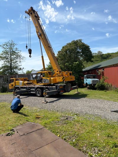 A yellow XCMG XCT20 crane is situated on a gravel and grassy area, with its boom extended towards the sky with clouds. A man wearing a blue shirt is visible near the base of the crane, facing a rusty metal plate on the ground. In the background, there are trees, a red barn-like structure, and a light blue truck. The setting appears to be outdoors, possibly a rural or industrial area. While the location is given as Kapreshumi, Georgia, there is no specific evidence of the city or landmarks to confirm it in the image.