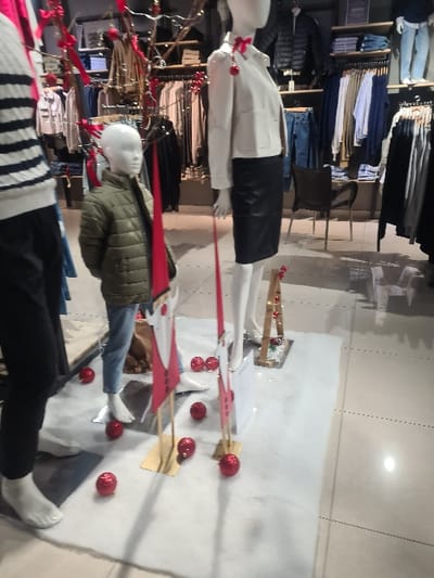 A store display features mannequins adorned with clothing. One mannequin wears a white jacket and black skirt, another a green jacket and jeans, and a third is dressed in a black and white striped top and black pants. The display incorporates red Christmas ornaments scattered on a snowy surface and branches with red bows, creating a festive ambiance. Garments on racks are visible in the background.