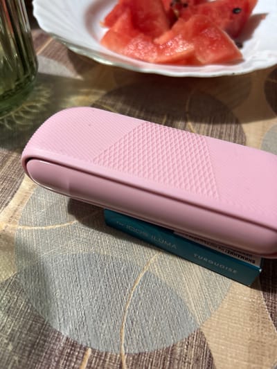 A pink protective case for an electronic device rests on a dark teal packaging of what appears to be tobacco sticks, likely "IQOS ILUMA Turquoise." The packaging is partially obscured by the pink case. In the background, a white plate holds several slices of watermelon. To the left, a green glass bottle or vase is visible, partially filled with a green liquid. The objects are placed on a patterned tablecloth with a textured surface. The scene is lit by overhead lighting, casting soft shadows. The location of this image is the city of Byala Slatina, Bulgaria.