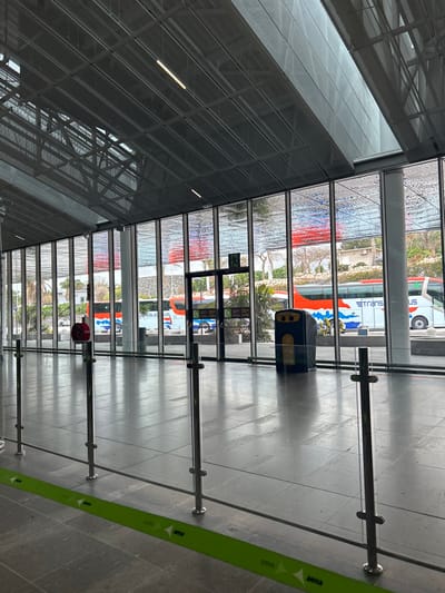The image shows an indoor space with a glass wall overlooking a paved outdoor area. There are four large buses parked outside, one of which is partially visible with the words "trans" and "us" on the side. The indoor space has a grey tiled floor and is enclosed by a stainless steel railing. A yellow and black recycling bin sits by the window. There is a green "atenza" sign on the floor. The image appears to have been taken from inside the Granadilla Airport in Tenerife, Spain, at the location defined by latitude 28.047 and longitude -16.58.