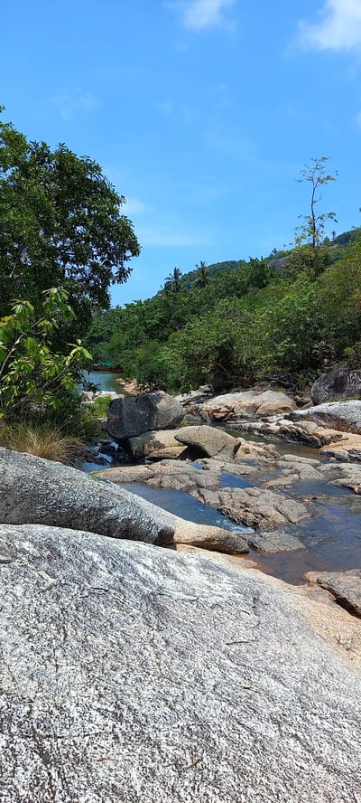 The image captures a vibrant, sunlit natural landscape, rotated 90 degrees clockwise. When viewed correctly (rotated counter-clockwise), it depicts a rocky stream or small waterfall winding through a lush, forested environment under a bright blue sky.
In the foreground, large, light-grey and brownish-grey granite-like rocks dominate the right and center, some showing water flowing over their textured surfaces or collecting in small, clear pools. The rock surfaces are rough and patterned. To the left, dense green foliage from trees and bushes frames the lower and middle sections of the scene.
The midground reveals a series of rocky cascades and pools, with a visible stream flowing downwards. The water appears clear, reflecting the blue sky in places. More large boulders and rocks are scattered along the stream banks, interspersed with green vegetation.
In the background, a heavily forested hillside or small mountain rises, covered in a canopy of rich green trees. A few distinct palm trees are visible amidst the dense foliage, characteristic of a tropical setting. The sky above is a clear, bright blue with a few scattered white, wispy clouds, indicating sunny weather. The bright lighting suggests it is daytime, likely mid-morning or afternoon.
No people, artificial objects, or visible text are present in the image. The scene conveys a sense of untouched natural beauty and tranquility, typical of a jungle or mountainous area with a freshwater source in Ban Tai, Thailand.