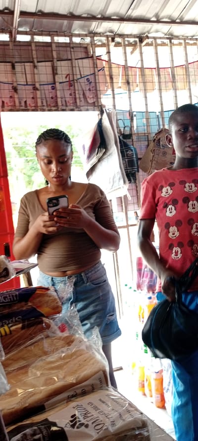 The image shows two young people standing in a market stall in Kwachiri, Nigeria. On the left, a young woman with braided hair is looking down at her phone, which she holds in both hands. She is wearing a brown t-shirt and denim shorts. In front of her, there are several loaves of bread wrapped in plastic. To the right of the woman, a young person wearing a red t-shirt with a Mickey Mouse pattern is holding a black plastic bag. The stall has a corrugated metal roof and shelves stocked with various items, including bags and bottles. The scene appears to be outdoors, with natural light filtering in, suggesting it is daytime. The overall atmosphere is casual and everyday. There are no clear indications of the weather or specific emotions, beyond the focused attention of the woman on her phone. Visible text on the bread packaging includes "Bon Pride Buttered Bread" and "Spec".