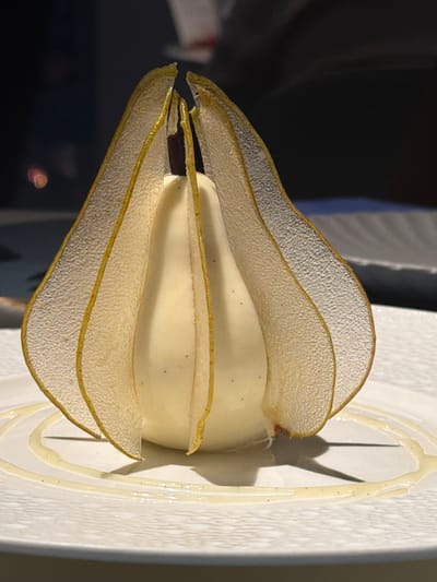A close-up, eye-level, medium shot captures a delicate dessert presented on a white plate in what appears to be a restaurant setting in Jianye District, China. The dessert is a white, dome-shaped confection, possibly ice cream or mousse, with a dark sliver protruding from its top. Encasing the confection are translucent, fan-shaped garnishes, likely made from thinly sliced fruit or pastry, with a golden-yellow edge. These garnishes are arranged to resemble petals, opening upwards and outwards from the dessert. The plate itself has a textured, embossed pattern and is adorned with a swirling drizzle of a light-colored sauce. The background is dark and softly blurred, with hints of colored lights, suggesting an indoor, possibly low-lit environment. The focus is sharply on the dessert and its immediate surroundings on the plate.