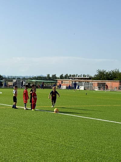 The image depicts a children's soccer practice on an artificial turf field with a light blue sky and scattered clouds. Several young boys in soccer uniforms are on the field. In the foreground, on the left, three boys stand in a line, one in a white and black striped jersey, another in an orange jersey, and a third in a red jersey. To their right, a boy in a dark blue and red jersey is running. A soccer ball is visible near the boy in the dark blue and red jersey. In the background, more children and adults are present on the field, some engaged in practice and others observing. A building with a red brick facade and white trim is situated behind the field, with trees and vegetation surrounding it. The field has white and yellow markings. Based on the context provided, this scene is located in Vidreres, Spain.