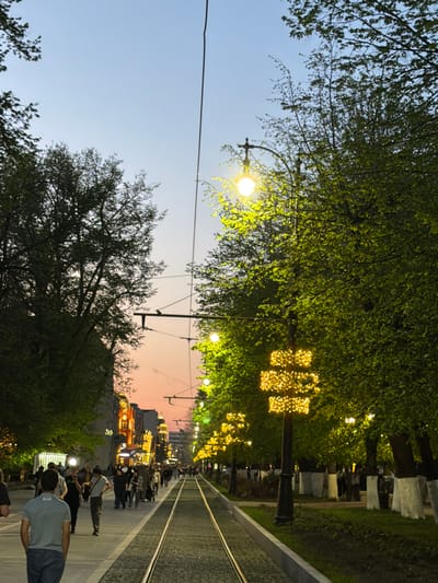 The image shows a street scene in Chermen, Russia, at dusk. A tram line runs down the center of the street, flanked by trees and sidewalks. String lights decorate the trees, and streetlights illuminate the scene. Pedestrians are walking along the sidewalks. The sky is transitioning from daylight to twilight, with a gradient of colors visible. Buildings are visible in the distance, lit up with lights.