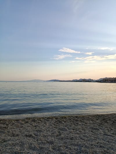 The image shows a beach scene in Águilas, Spain. The foreground consists of a gravel beach. Gentle waves lap the shore. In the middle ground is calm, flat water. In the background, there is a skyline of buildings, hills, and distant mountains. The sky is pale blue with some light clouds near the horizon. The light indicates that it is either sunrise or sunset. There are no visible people in the scene.