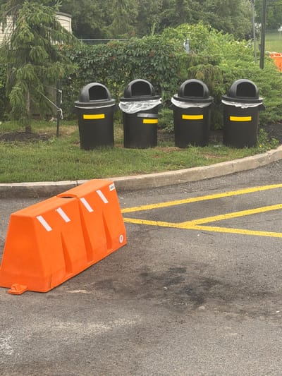 Four black trash cans with yellow bands are lined up on a grassy area behind a low concrete curb. The trash cans are partially covered with plastic liners, with one can showing a more significant amount of plastic liner. An orange plastic barrier is in the foreground, placed on an asphalt surface marked with yellow lines. The barrier is angled and positioned to the left of the frame. Behind the trash cans, a dense hedge and trees form a natural backdrop. A thin, wire-like object is visible extending from the grassy area behind the trash cans, secured to a stake. The scene appears to be outdoors, possibly in a parking lot or public area.