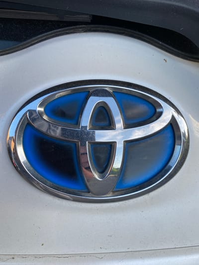 The image is a close-up, eye-level shot of a car emblem. The emblem is oval-shaped with a chrome border and a blue interior. Inside the blue area, there is a stylized, metallic silver design that resembles an abstract representation of overlapping curves or loops. The emblem appears to be mounted on a white car body, which has a slightly textured surface and is showing signs of dust and dirt. To the left of the emblem, a portion of a black, textured car part is visible, possibly a fender or trim piece. The lighting suggests it is daytime, likely outdoors, given the bright illumination and reflections on the chrome. No people or specific activities are depicted. There is no visible text in the image. The location context of Lütjenwestedt, Germany, is provided, but there are no specific visual cues in the image to confirm this location.