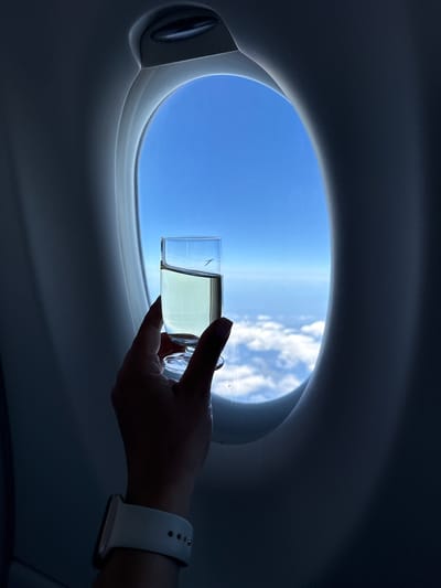 The image shows a hand holding a glass of clear liquid with a white smartwatch on the wrist, in front of an airplane window. Outside the window, there is a clear blue sky with some white clouds below. The interior of the airplane cabin is dark and out of focus, framing the window. There is no discernible indication of the city of Rudeni, Romania.