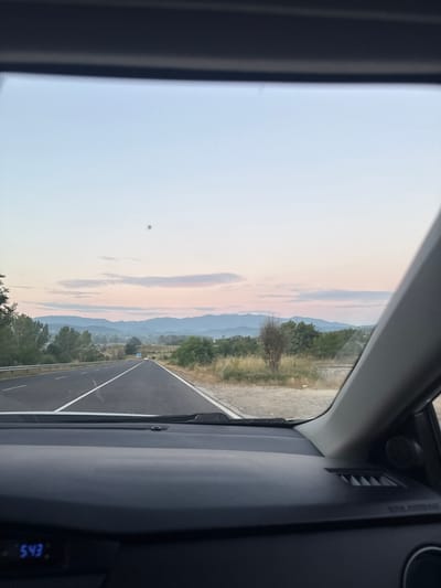 The image shows a view from inside a vehicle looking out the windshield. The scene depicts a road stretching into the distance, flanked by trees and vegetation. In the background, a range of mountains can be seen under a sky with soft, pastel-colored clouds, suggesting it is either sunrise or sunset. The road itself appears to be a highway with white lane markings. The interior of the vehicle is visible on the right side of the frame, including parts of the dashboard and a digital display showing "5:43". The overall impression is of a scenic drive during twilight hours in a rural or mountainous region. There is some text visible on the car's interior, possibly an airbag warning, but it is difficult to read clearly.