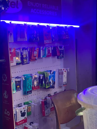 This media file captures a display of mobile phone accessories in a retail setting. The scene is brightly lit by what appears to be a blue LED light strip running vertically along the left side of the image, casting a vibrant glow. The accessories are neatly arranged on pegboards, showcasing a variety of items such as data cables, batteries, earphones, and what seem to be wireless earbuds. The packaging for these items is colorful and varied, with brands like "LINNCO" and "G-POWER" visible.
Text overlays on the left side of the image include "el", "ENJOY RELIABLE", and "ACCESSORIES", indicating the store's branding. Along the bottom of the image, more text suggests a product's origin and quality, with phrases like "Durable Material", "Sold in 50+ Countries", and "Professional Tested". A small graphic of the globe is also present, reinforcing the international aspect of the sales.
The overall impression is of a well-stocked shop aiming to attract customers with a diverse range of mobile accessories. The blue lighting creates a modern and energetic atmosphere. The image seems to be taken at a point of sale within the store, possibly in Barakin, Nigeria, as indicated by the location context. There are no people visible in the image, emphasizing the focus on the products themselves.