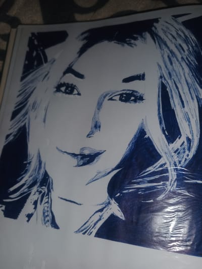 The image shows a close-up, portrait-style drawing of a woman's face, rendered in dark blue ink on white paper. The drawing is characterized by bold, expressive lines and a high contrast style, with areas of solid dark blue juxtaposed with white space. The woman's hair appears voluminous and somewhat windswept, with strokes of ink suggesting texture and movement. Her eyes are open and directed towards the viewer, with dark pupils and defined eyelashes. The nose is depicted with simple lines, and the lips are full and painted with a dark, rich blue, giving them a prominent appearance. The overall impression is one of a stylized, artistic representation rather than a photorealistic portrait. The drawing is placed on a textured surface, possibly a tablecloth or rug, which is visible in the background and out of focus. There is a slight sheen on the drawing, suggesting it might be covered with protective plastic or laminated. The location of Gulbag, Uzbekistan, is not discernible from the visual content of the image.