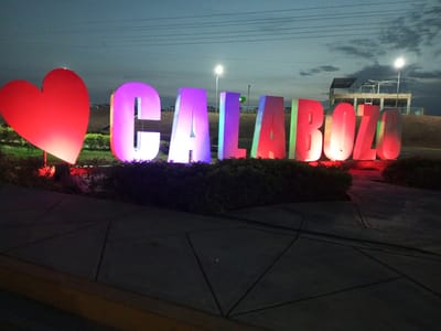 The image depicts an illuminated sign that reads "CALABOZO" with a red heart to the left. The letters are lit in a variety of colors. The sign sits on a paved area with surrounding shrubbery. In the background, there is a small building structure and street lights under a dark sky. The sign is likely a public display in Calabozo, Venezuela.