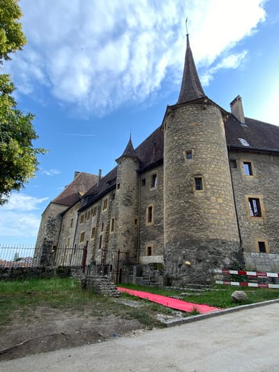 In the city of Milvignes, Switzerland, a stone castle with a conical turret and a tall chimney stands under a bright blue sky with scattered clouds. A red carpet is laid out on the grass leading to the castle entrance. The castle is made of rough-hewn stones and has numerous windows. A metal fence runs along the side of the castle. Several red and white barricades with "DUCKERT" printed on them are visible in front of the castle. A large rock sits near the barricades. The ground in front of the castle is a dirt path with tire tracks. Lush green grass and trees are present around the castle.
