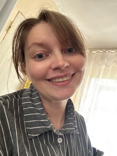 A close-up, low-angle portrait shows a young person with fair skin, light brown hair, and blue eyes, smiling broadly directly at the camera. Their head is tilted, and a bright, genuine smile reveals their teeth. They are wearing a grey collared shirt with thin white vertical stripes, and a single white button is visible on the placket. The background is brightly lit, featuring a window with ornate white lace curtains that filter the natural light. Above the window, a light-colored wall or ceiling is visible, possibly adorned with decorative white molding or a cornice. The bright lighting suggests it is daytime. The overall mood conveyed by the subject's expression is happy and friendly. The scene is indoors, likely within a home, located in Aksay, Kazakhstan. No text is visible in the image.