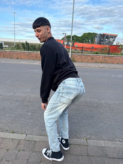 In Boston, United Kingdom, a young man bends forward, looking over his right shoulder with a slight smile. He has short, dark hair with a distinct fringe and faded sides, light facial stubble, and a prominent nose. He is dressed in a plain black crew-neck sweatshirt, light-wash denim jeans revealing a black underwear waistband with partial white text, and black canvas high-top sneakers with white laces and a star logo on the ankle. He stands on a grey paved sidewalk next to an asphalt road with faint white markings. A red brick wall topped with a black metal fence spans the midground. Behind the fence, a large, weathered red and black structure resembling a ship's hull and superstructure is prominently visible, bearing the inscription 'LO. 243'. Green trees and a distant light-colored building with a white roof are in the background, beneath a partly cloudy blue sky. A tall grey street lamp pole is positioned on the left side of the frame.