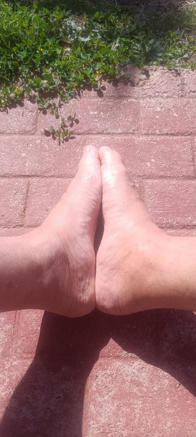 A pair of bare human feet are centrally positioned on a reddish-brown paved surface, oriented vertically with toes upward. The feet are placed with their soles touching and heels slightly apart. Visible skin is light-toned with minor hair growth on the dorsal surfaces. Distinct shadows from the feet are cast onto the pavement, indicating direct sunlight from a moderate angle. The upper portion of the frame displays green ground cover vegetation, consisting of small-leaved plants and occasional tiny yellow flowers, bordering the paved area. The image is captured from an overhead perspective at Forest Hills, Spain.