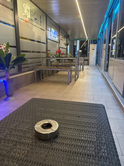 The image depicts an outdoor seating area, likely a patio or terrace, of a establishment in Okop, Bulgaria. The foreground features a dark woven table with a metal ashtray placed on its surface. The table has a textured, patterned finish. Beyond the table, the scene extends to reveal a row of outdoor tables and benches, suggesting a dining or waiting area. These tables have metal frames and wooden slats. Plants are visible, including a potted plant with large green leaves and another with red flowers. The backdrop consists of a building with large glass windows and frosted glass panels, some of which are illuminated with blue LED lighting along the top. A white vertical appliance, possibly an air conditioner or heater, is situated near the windows. The flooring is tiled with large, light-colored rectangular tiles. The overall ambiance suggests a commercial establishment with outdoor seating.
