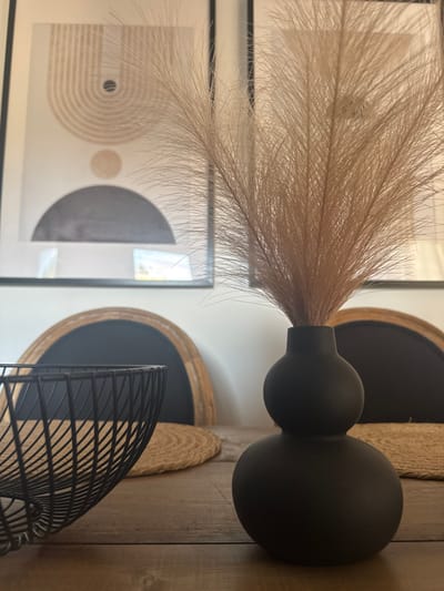 The image displays an interior still life arrangement on a wooden table. In the foreground to the right, a matte black, double-gourd-shaped vase holds a bundle of light brown, feathery pampas grass. To the left, a black metal wire bowl is partially visible. Both objects rest on a light brown, rustic wooden table surface. Two circular placemats woven from a natural fiber, possibly jute, are positioned on the table, one beneath the wire bowl and another partially obscured behind the vase.

In the mid-ground, behind the table, two chairs are visible. Each chair features a dark, semicircular back framed by a lighter, possibly wooden or rattan border. On the light-colored wall above the chairs, two framed abstract art prints are hung. The left print prominently features geometric shapes including concentric arcs and circles in beige and dark gray tones on a white background. The right print is largely obscured by the pampas grass. The scene depicts a modern, minimalist interior. No individuals are present. The image provides no visual evidence to confirm a specific location within the city of La Pedrera, Spain.