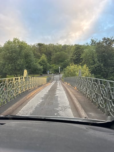 The image shows a view from a vehicle driving across a bridge. The bridge has ornate metal railings on both sides, with a circular pattern. The road surface of the bridge is wet, indicating recent rain or dew. Lush green trees line the sides of the bridge, suggesting a natural, possibly rural, setting. In the distance, there are signs, one of which appears to be a directional sign with a blue arrow and a white icon. The sky is partly cloudy, with patches of sunlight breaking through, suggesting it is either early morning or late afternoon. The overall impression is one of a peaceful, perhaps slightly overcast, day. There is no discernible text on the railings or the road, though some faint markings are visible on the wet surface. The scene evokes a sense of travel and transition. The location context provided is Telford, United Kingdom.