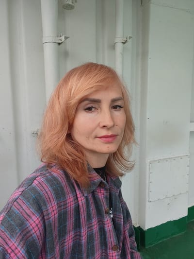 A woman with peach-colored hair and a pink and gray plaid shirt is photographed from the chest up, looking towards the camera. She is standing in front of a white wall with vertical pipes. A green stripe runs along the bottom of the wall. The image appears to have been taken outdoors, possibly on a boat or ferry, given the industrial-looking wall and railing. The location is identified as Topornya, Russia.