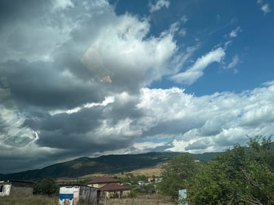 This image, taken in Ismailli District, Azerbaijan, presents a landscape with a dramatic sky reflected on a surface, likely a car windshield. The scene is captured in an unusual orientation, with the sky appearing at the bottom and the ground at the top.
The reflected elements at the top of the image show a rural setting. Trees with green foliage and hints of red berries are visible, along with buildings that appear to be houses and some with corrugated metal roofs. Beyond the immediate structures, a valley with dark, rolling hills stretches into the distance under a vast sky. The sky itself is filled with a mix of light, fluffy white clouds and darker, more ominous storm clouds. This suggests a dynamic weather pattern, possibly transitioning from fair to stormy or vice versa.
The reflection on the surface below creates a surreal effect. The clouds dominate this lower portion of the image, with a bright blue sky peeking through in some areas. Within the clouds, there are faint reflections of what appear to be human hands and possibly the upper body of a person, creating an ethereal or ghostly impression. A light-colored car, likely the vehicle from which the photo was taken, is also partially visible in the reflection. The combination of the natural landscape and the reflected elements creates a sense of depth and visual intrigue. The time of day is not explicitly clear but the lighting suggests it could be late morning or afternoon.