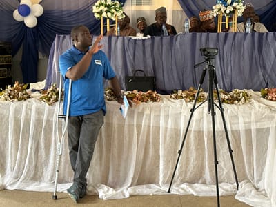 An indoor event, likely a meeting or presentation, is captured in Unguwan Dosa, Nigeria. In the foreground, a male speaker stands prominently, facing right and gesturing with his right hand. He wears a blue polo shirt with the text "NDI" on the chest, gray trousers, and sneakers. He leans on a silver crutch, visible under his left arm, and holds a small notepad or book in his left hand.

Behind a long table draped in white and blue fabric, several men are seated, partially visible from the chest up. They wear traditional caps and attire, with some appearing attentive to the speaker, while others are seen drinking from plastic water bottles. The table is decorated with artificial floral arrangements featuring white, blue, and brown flowers. A black briefcase or laptop bag rests on the table in front of the seated panel.

To the right, a black video camera mounted on a tripod is positioned, seemingly recording the proceedings. The background features blue and white fabric draping, with a decorative cluster of white and blue balloons arranged like a flower on the left. The lighting suggests the event takes place during the daytime.